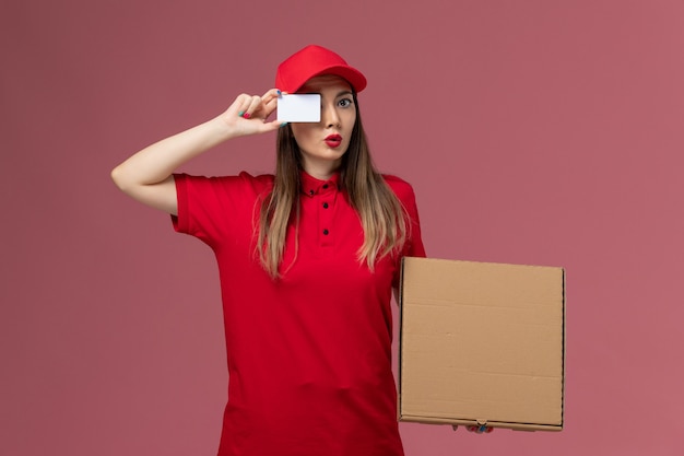 Front view young female courier in red uniform holding delivery food box and white card on light-pink background delivery service uniform company job