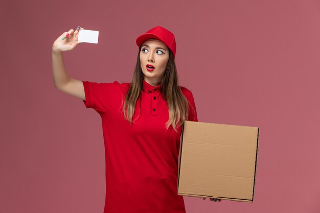 Front view young female courier in red uniform holding delivery food box and white card on the pink background delivery service uniform company job