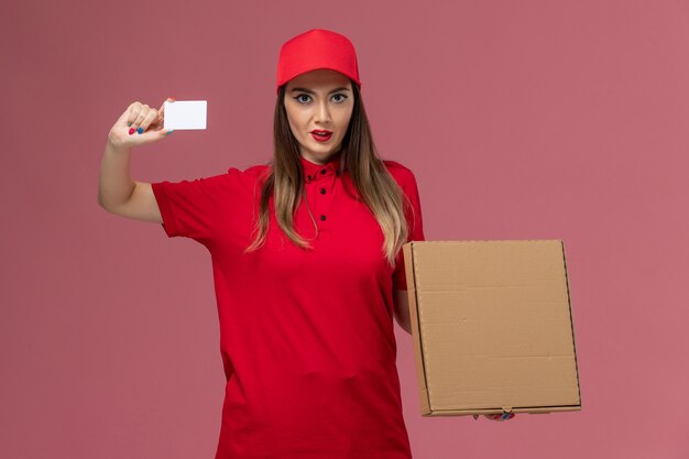 Front view young female courier in red uniform holding delivery food box and white card on the pink background delivery service uniform company