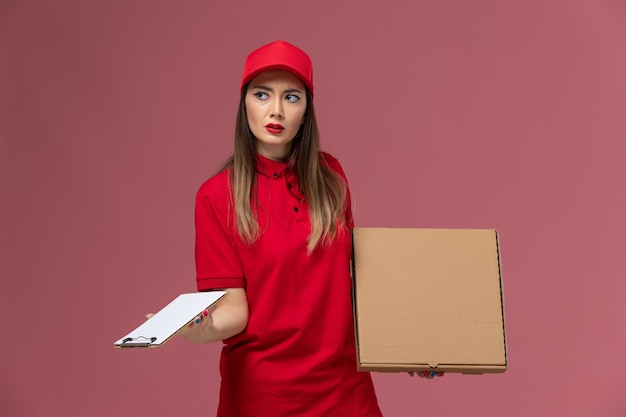 明るいピンクの背景にメモ帳付きの配達フードボックスを保持している赤い制服の正面図若い女性の宅配便配達サービス制服会社の仕事