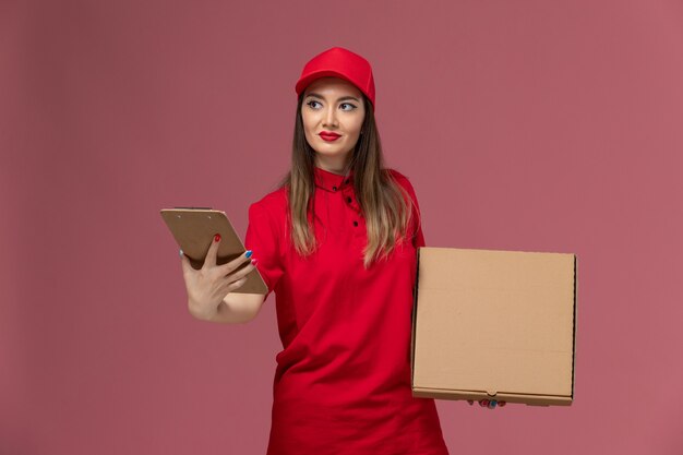 Вид спереди молодая женщина-курьер в красной форме, держащая коробку с едой для доставки с блокнотом на светло-розовом фоне, служба доставки униформа компании