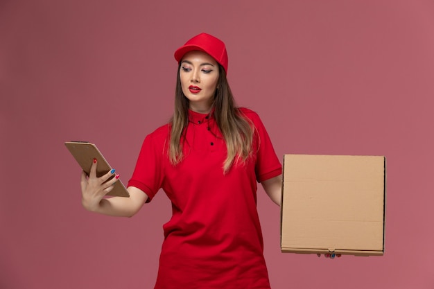 Front view young female courier in red uniform holding delivery food box with notepad on pink floor delivery service uniform company job