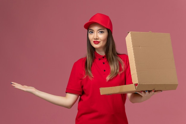 Front view young female courier in red uniform holding delivery food box with slight msile on light-pink background service delivery job uniform