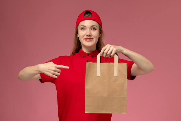 淡いピンクの壁に配達紙食品パッケージを保持している赤い制服を着た若い女性の宅配便の正面図