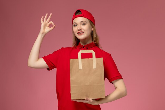 Front view young female courier in red uniform holding delivery paper food package on light-pink wall