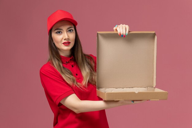 明るいピンクの背景に空のフードボックスを保持している赤い制服を着た若い女性の宅配便の正面図