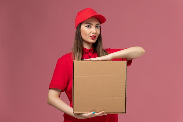 Front view young female courier in red uniform holding food box on the pink background worker service delivery uniform company job