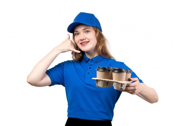 Вид спереди молодой женщины-курьера в униформе, держащей чашки с кофе, улыбаясь, показывая позу телефонного звонка