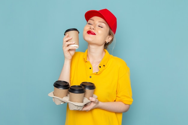 Front view young female courier in yellow shirt and red cape holding coffee cups on the blue space  work 
