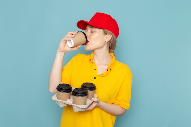 Молодая женщина-курьер в желтой рубашке и красной накидке, держащая кофейные чашки, пьющая на синем космическом работнике, вид спереди