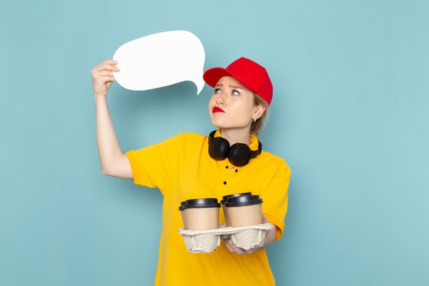 Front view young female courier in yellow shirt and red cape holding coffee cups and white sign on the blue space job work  