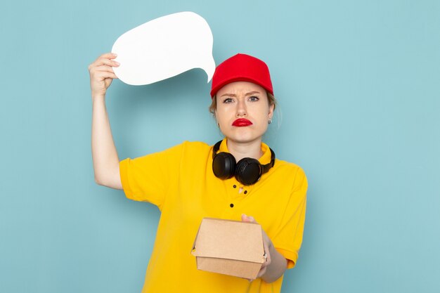 Front view young female courier in yellow shirt and red cape holding food package and white sign on the blue space   