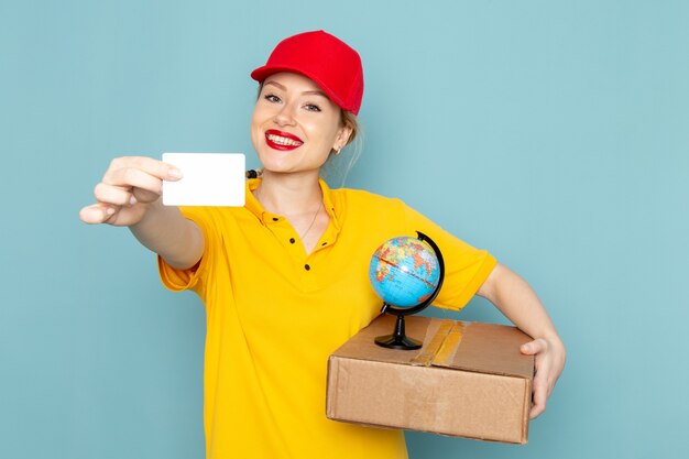 Front view young female courier in yellow shirt and red cape holding little globe and package smiling on the blue space   