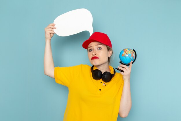 Front view young female courier in yellow shirt and red cape holding little globe and white sign with displeased expression on the blue space   