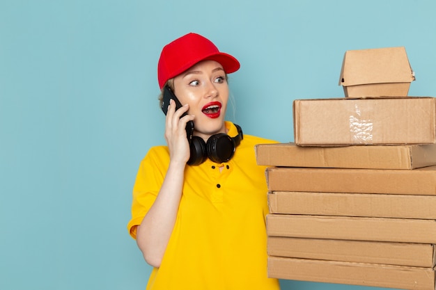 Front view young female courier in yellow shirt and red cape holding multiply  packages talkign on the phone on the blue space   job