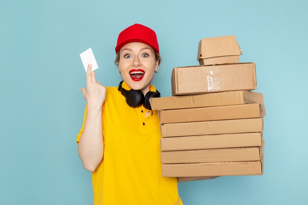 Front view young female courier in yellow shirt and red cape holding multiply  packages and white card on the blue space  