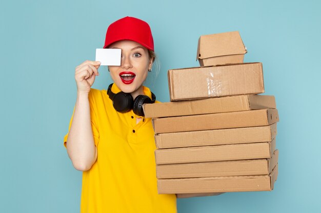 Front view young female courier in yellow shirt and red cape holding multiply  packages with card on the blue space   job