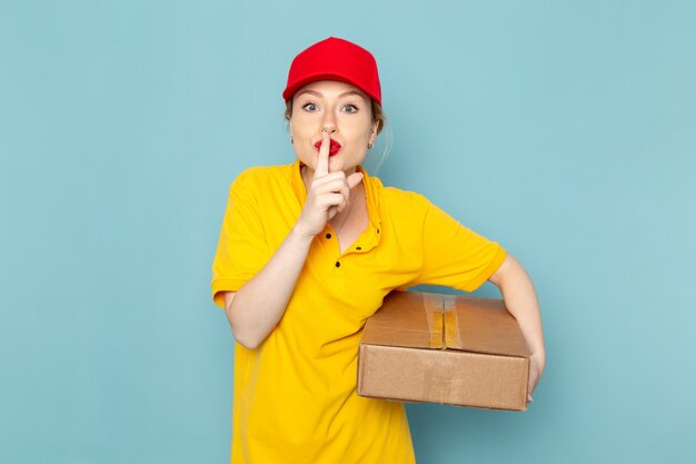 黄色のシャツと青い空間の仕事の仕事にパッケージを保持している赤いマントの正面の若い女性宅配便