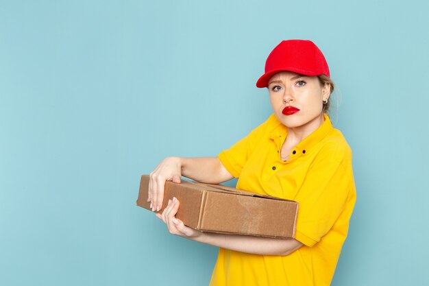 Front view young female courier in yellow shirt and red cape holding package on the blue space  