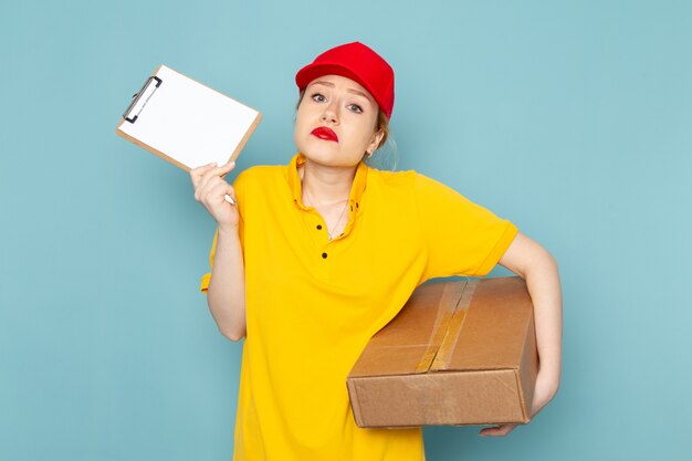 Front view young female courier in yellow shirt and red cape holding  package and notepad on the blue space  job 