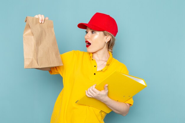 Front view young female courier in yellow shirt and red cape holding  package and yellow file on the blue space  work 