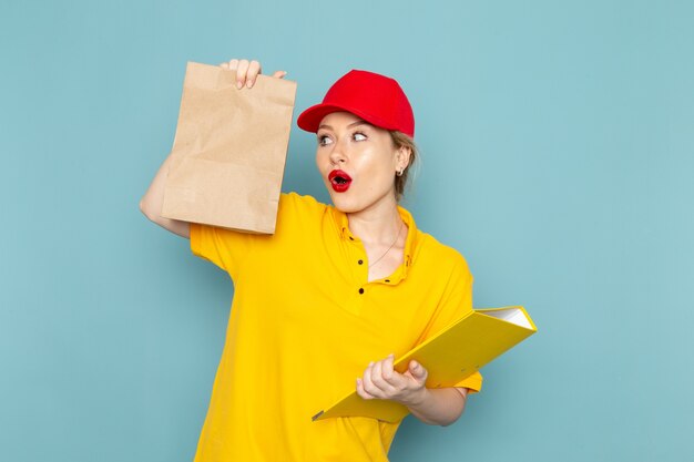 Front view young female courier in yellow shirt red cape holding  package and yellow file on the blue space  worker 