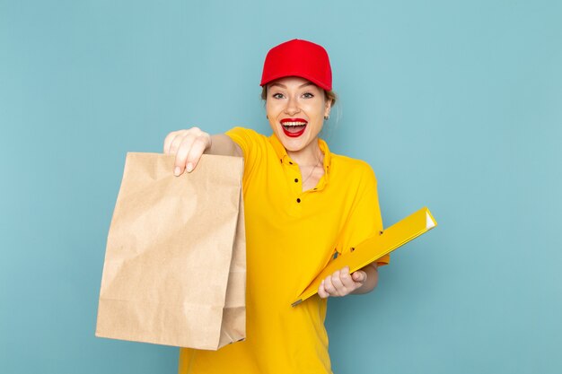 Front view young female courier in yellow shirt and red cape holding  package and yellow file on the blue space  