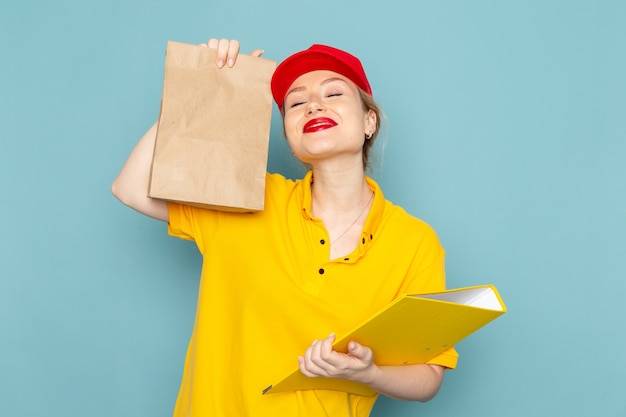 Front view young female courier in yellow shirt and red cape holding  package and yellow file smiling on the blue space  worker 