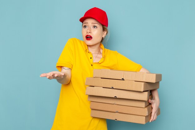 Front view young female courier in yellow shirt and red cape holding packages on the blue space worker