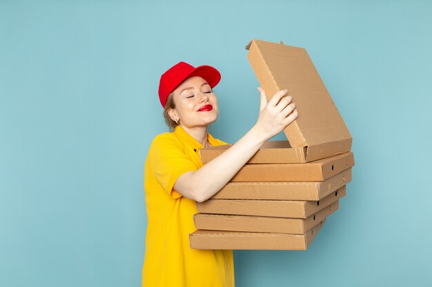 Front view young female courier in yellow shirt and red cape holding  packages opening them on the blue space  