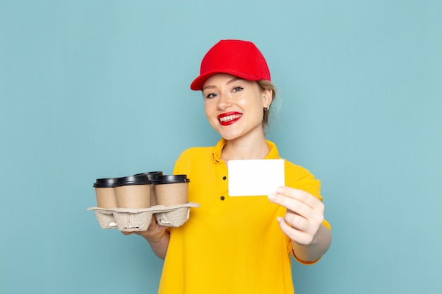 Foto gratuita giovane corriere femminile di vista frontale in camicia gialla e mantello rosso che tiene le tazze di caffè di plastica sul lavoro della ragazza della donna dello spazio blu