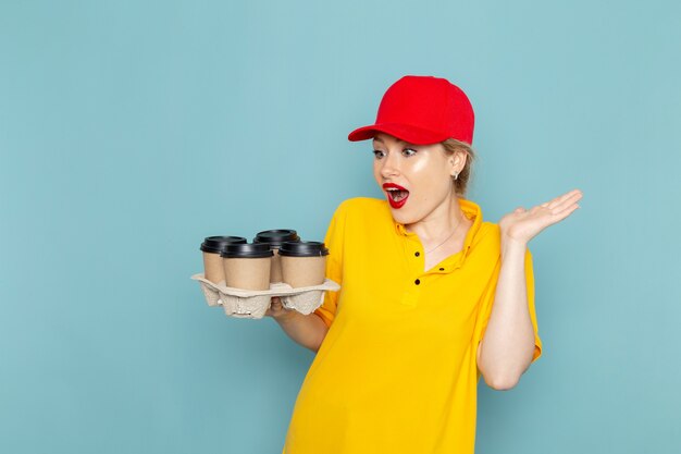 Front view young female courier in yellow shirt and red cape holding plastic coffee cups on the blue space   woman lady girl job