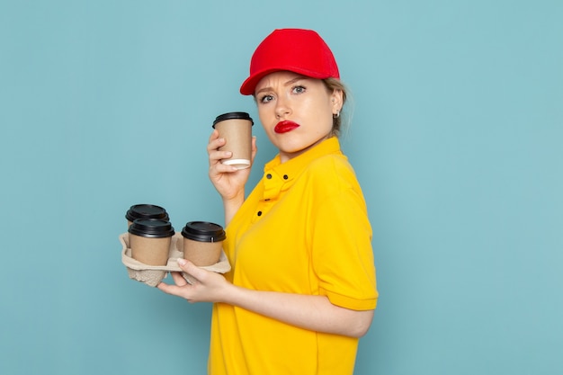 Front view young female courier in yellow shirt and red cape holding plastic coffee cups on the blue space  