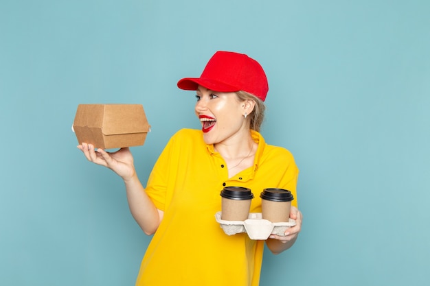 Front view young female courier in yellow shirt and red cape holding plastic coffee cups and food package on the blue space   job