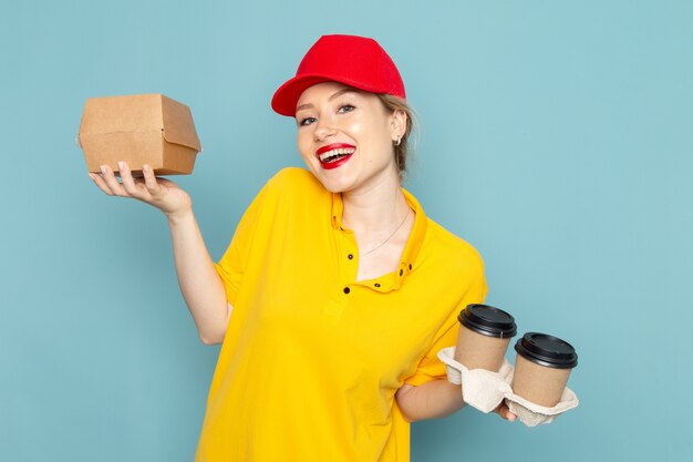 Front view young female courier in yellow shirt and red cape holding plastic coffee cups and food package on the blue space 