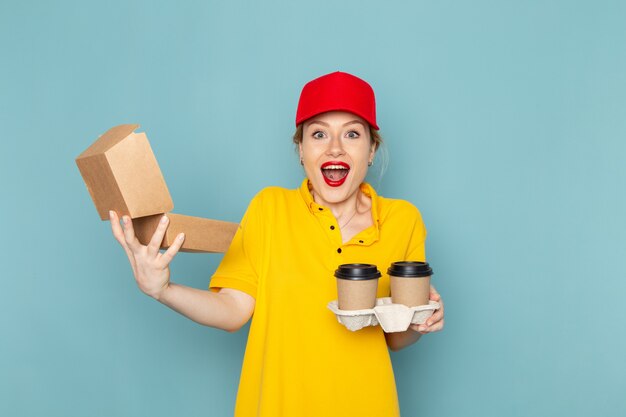Front view young female courier in yellow shirt and red cape holding plastic coffee cups and food package smiling on the blue space  job 
