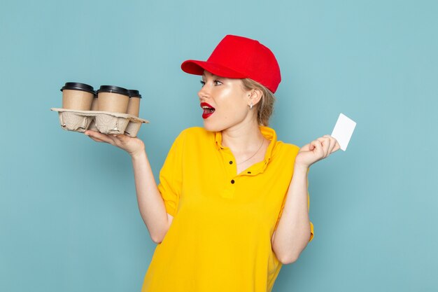 Front view young female courier in yellow shirt and red cape holding plastic coffee cups white card on the blue space   woman