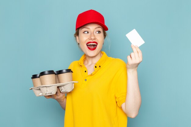 黄色のシャツと青いケープのプラスチック製のコーヒーカップの白いカードを保持している赤いマントの正面若い女性宅配便