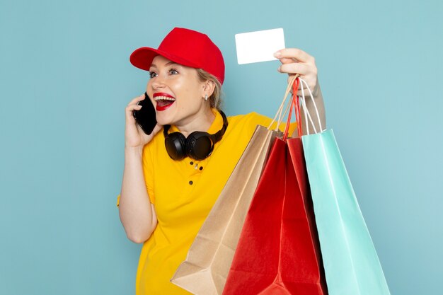 Front view young female courier in yellow shirt and red cape holding shopping packages card talking on the phone on the blue space   job