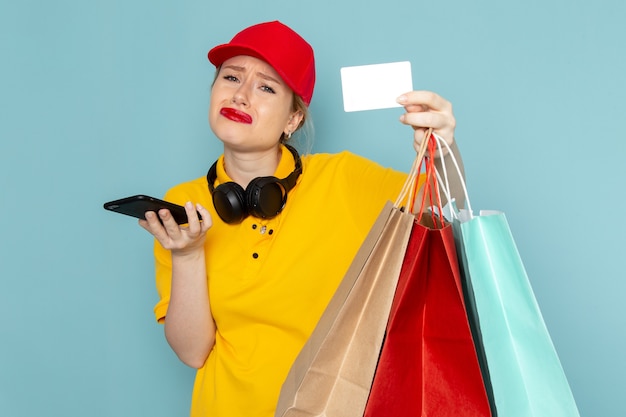 Front view young female courier in yellow shirt and red cape holding shopping packages phone and card on the blue space  
