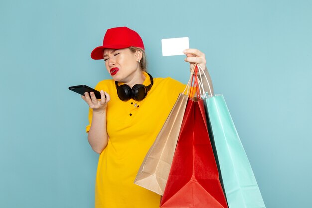 Молодая женщина-курьер в желтой рубашке и красной накидке, держащая пакеты с покупками, разговаривает по телефону на синем полу, вид спереди