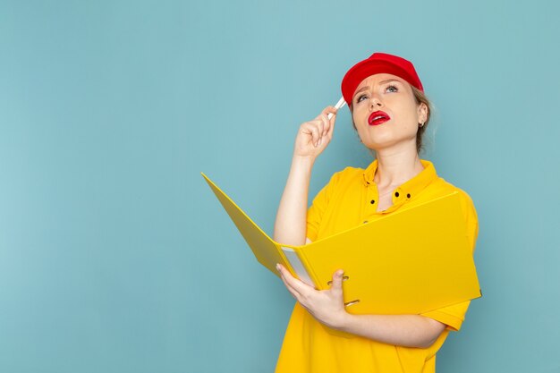 Front view young female courier in yellow shirt and red cape holding yellow file thinking on the blue space  worker 