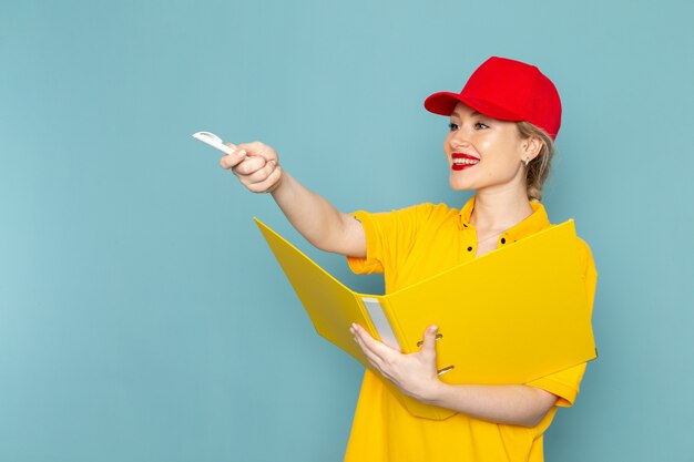 Front view young female courier in yellow shirt and red cape holding yellow file writing down notes on the blue space  job 