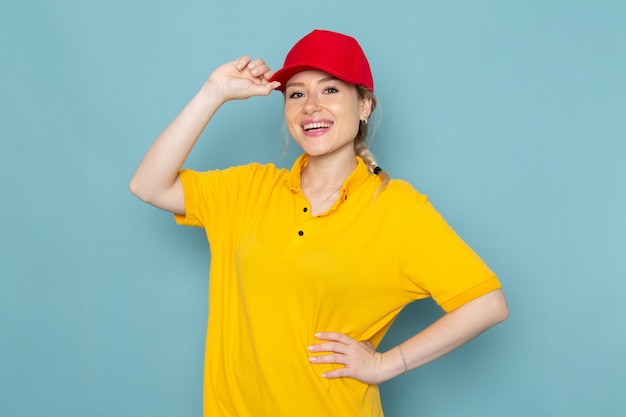 Вид спереди молодая женщина-курьер в желтой рубашке и красной накидке улыбается и позирует на синем космическом рабочем месте.