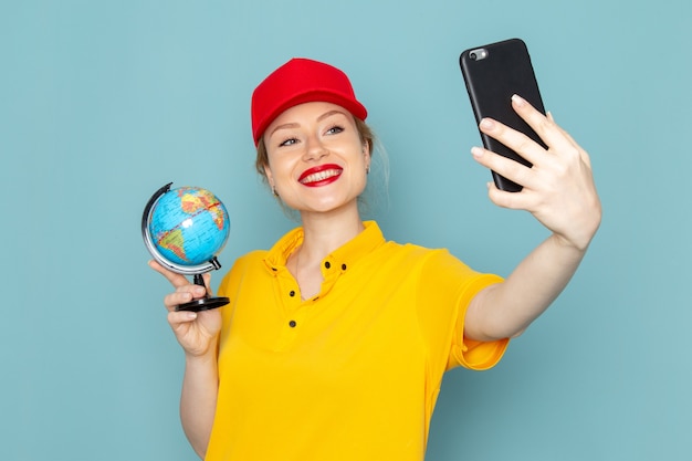 Front view young female courier in yellow shirt and red cape taking a selfie with globe on the blue space  