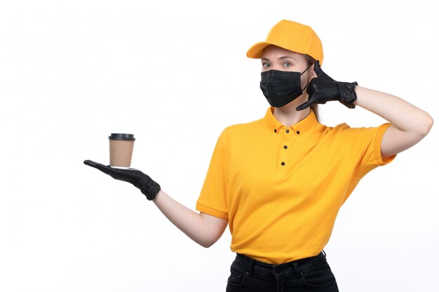 A front view young female courier in yellow uniform black gloves and black mask holding coffee cup asking for a call