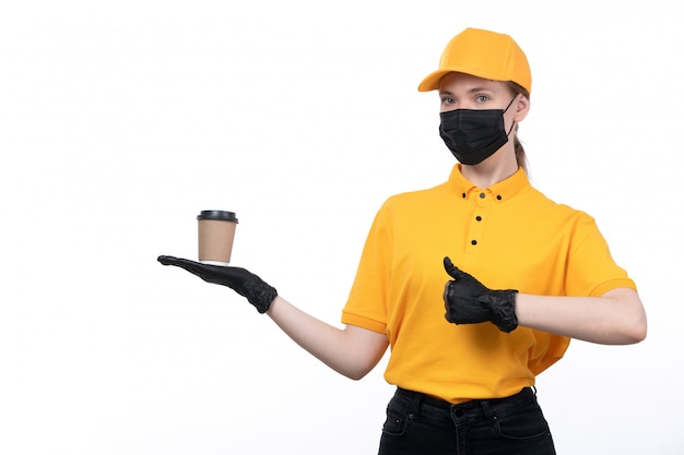 A front view young female courier in yellow uniform black gloves and black mask holding coffee cup showing like sign