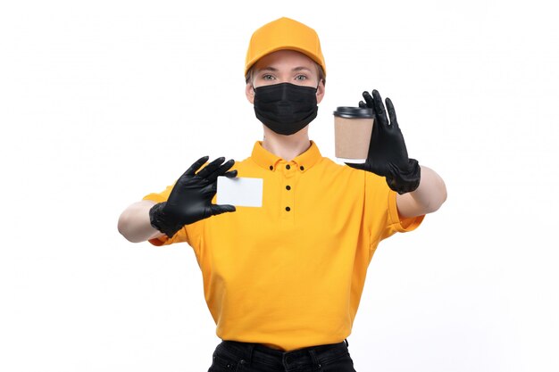 A front view young female courier in yellow uniform black gloves and black mask holding coffee cup and white card