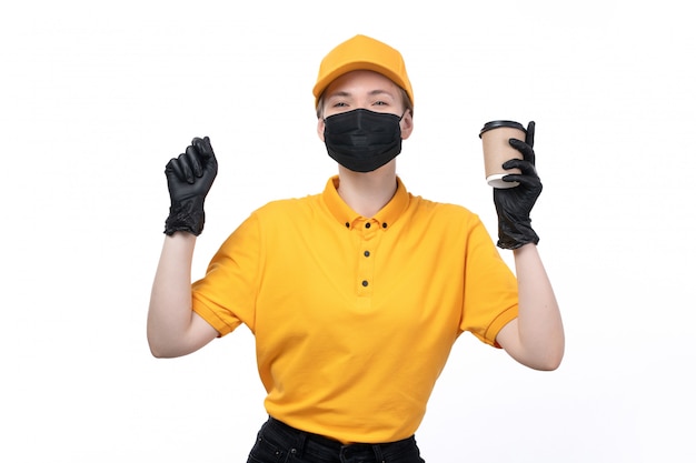 A front view young female courier in yellow uniform black gloves and black mask holding coffee cup with great mood