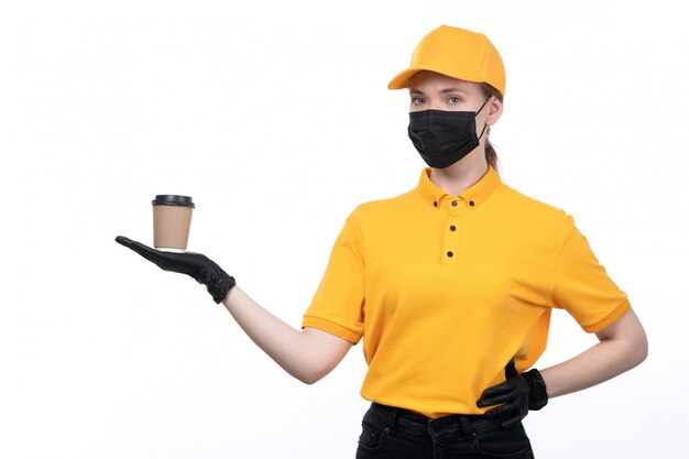 A front view young female courier in yellow uniform black gloves and black mask holding coffee cup